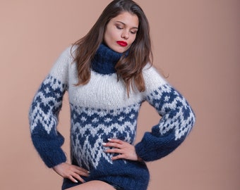 Puede incluir: Una mujer con un suéter azul y blanco con un cuello alto. Está sentada en un taburete de madera con las piernas cruzadas.