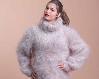 Puede incluir: Un suéter de cuello alto de color beige claro, grueso y de corte holgado, confeccionado con un tejido de punto esponjoso y de pelo largo. La prenda presenta mangas largas, un cuello acanalado grueso y un ajuste relajado.