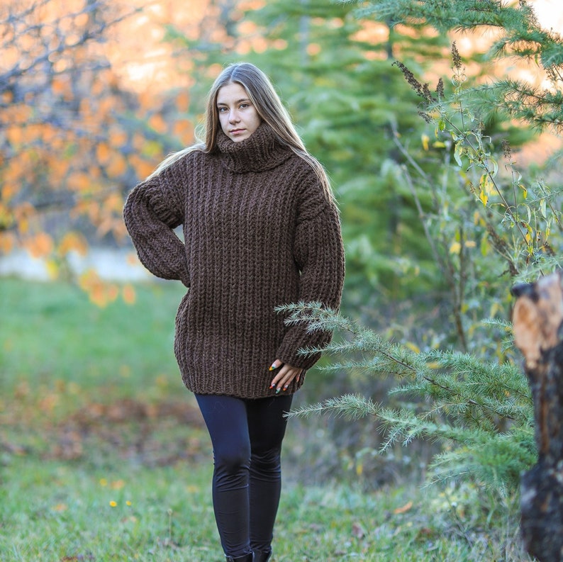 May include: A brown, oversized turtleneck sweater with a chunky knit design. The sweater is long-sleeved and worn with black leggings. The person is standing outdoors in a natural setting with green foliage and a blurred background.