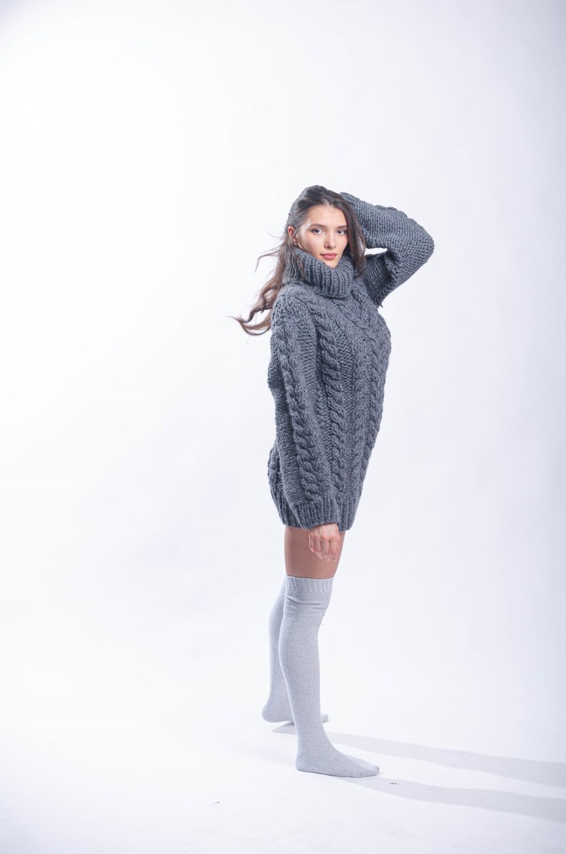 May include: A woman wearing a gray cable knit sweater and gray knee-high socks. She is standing on a white background.