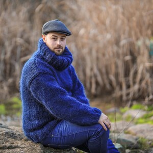 May include: A man wearing a blue turtleneck sweater and a dark gray newsboy cap sits on a rock. He is looking at the camera.