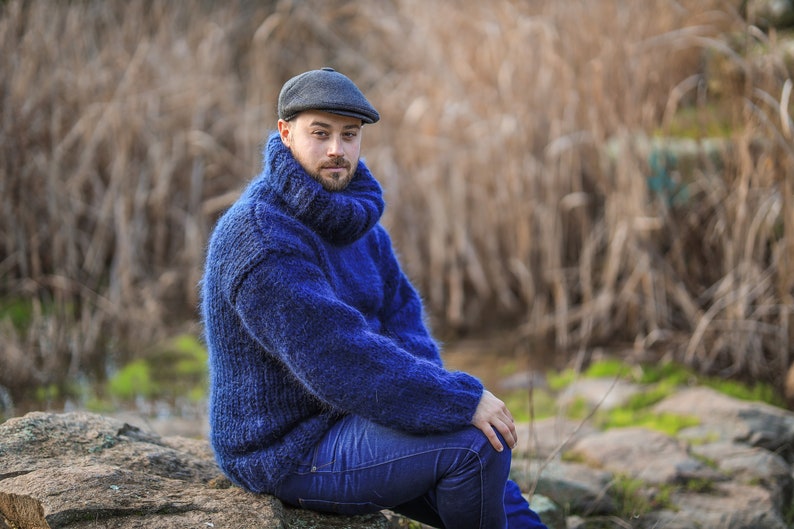 May include: A man wearing a blue turtleneck sweater and a dark gray newsboy cap sits on a rock. He is looking at the camera.