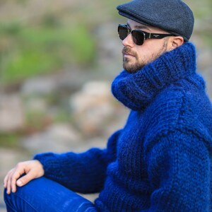 May include: A man wearing a blue turtleneck sweater, blue jeans, and a black newsboy cap. He is wearing sunglasses and has a beard.