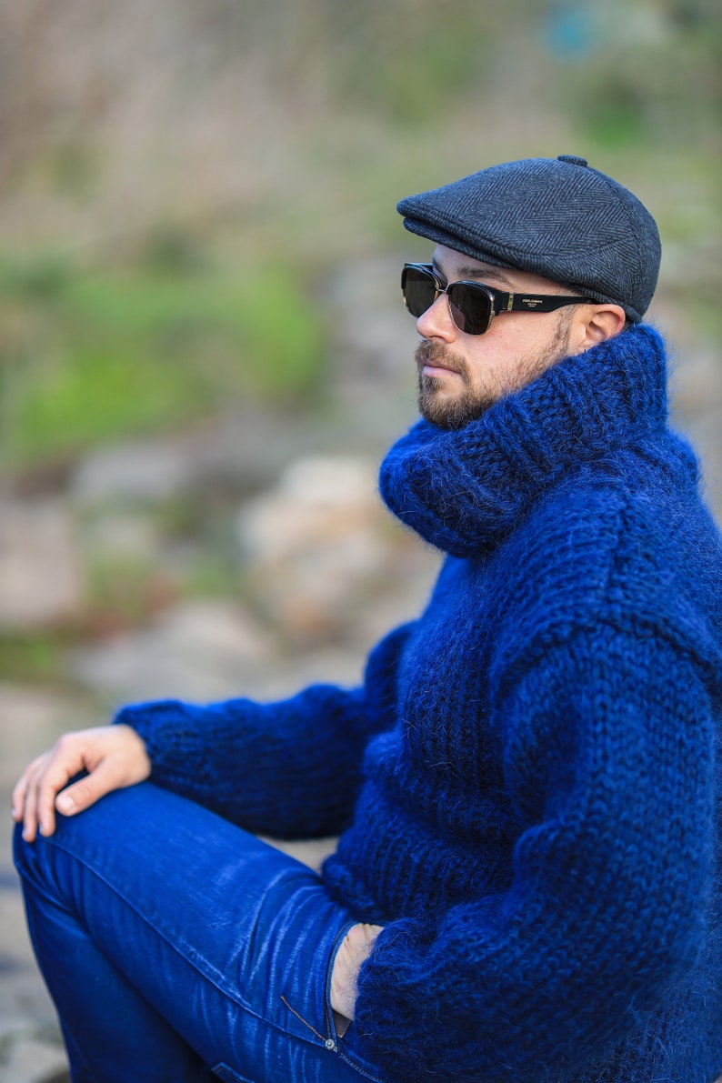 May include: A man wearing a blue turtleneck sweater, blue jeans, and a black newsboy cap. He is wearing sunglasses and has a beard.
