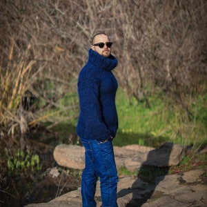 May include: A man wearing a blue turtleneck sweater and blue jeans stands on a rocky path. He is wearing sunglasses and brown shoes.