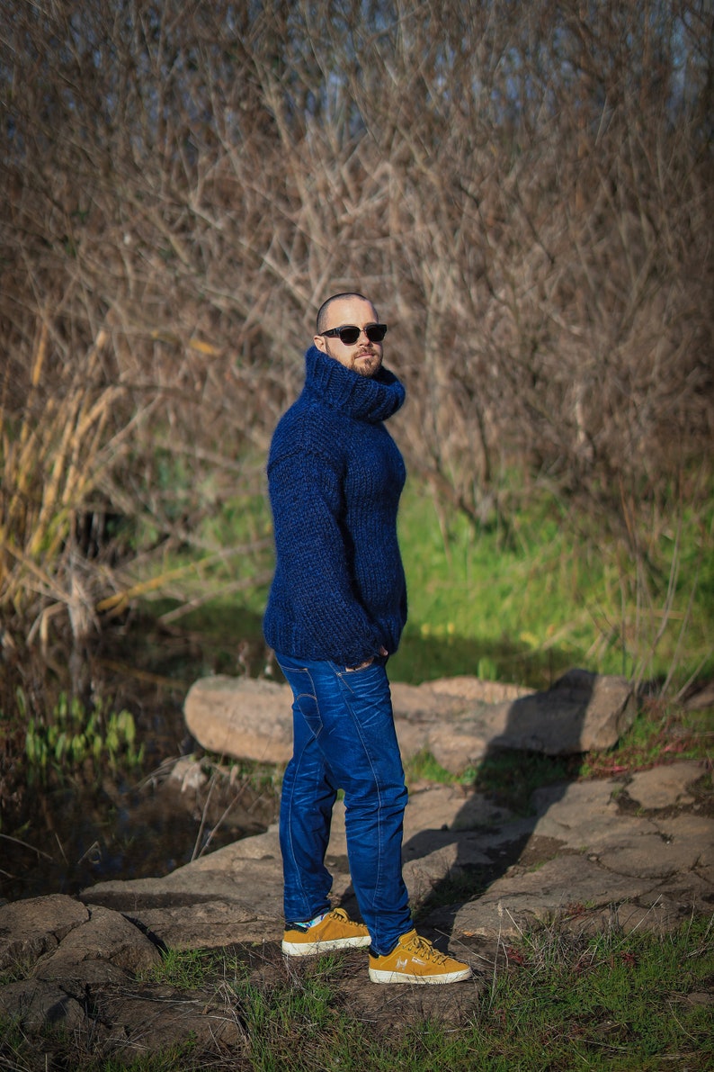 May include: A man wearing a blue turtleneck sweater and blue jeans stands on a rocky path. He is wearing sunglasses and brown shoes.