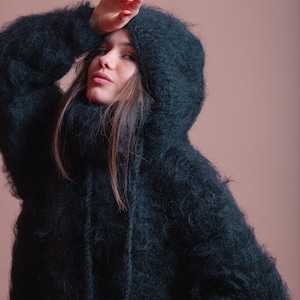 May include: A woman wearing a dark green fuzzy hooded sweater. The sweater has a high neck and a long, loose fit.