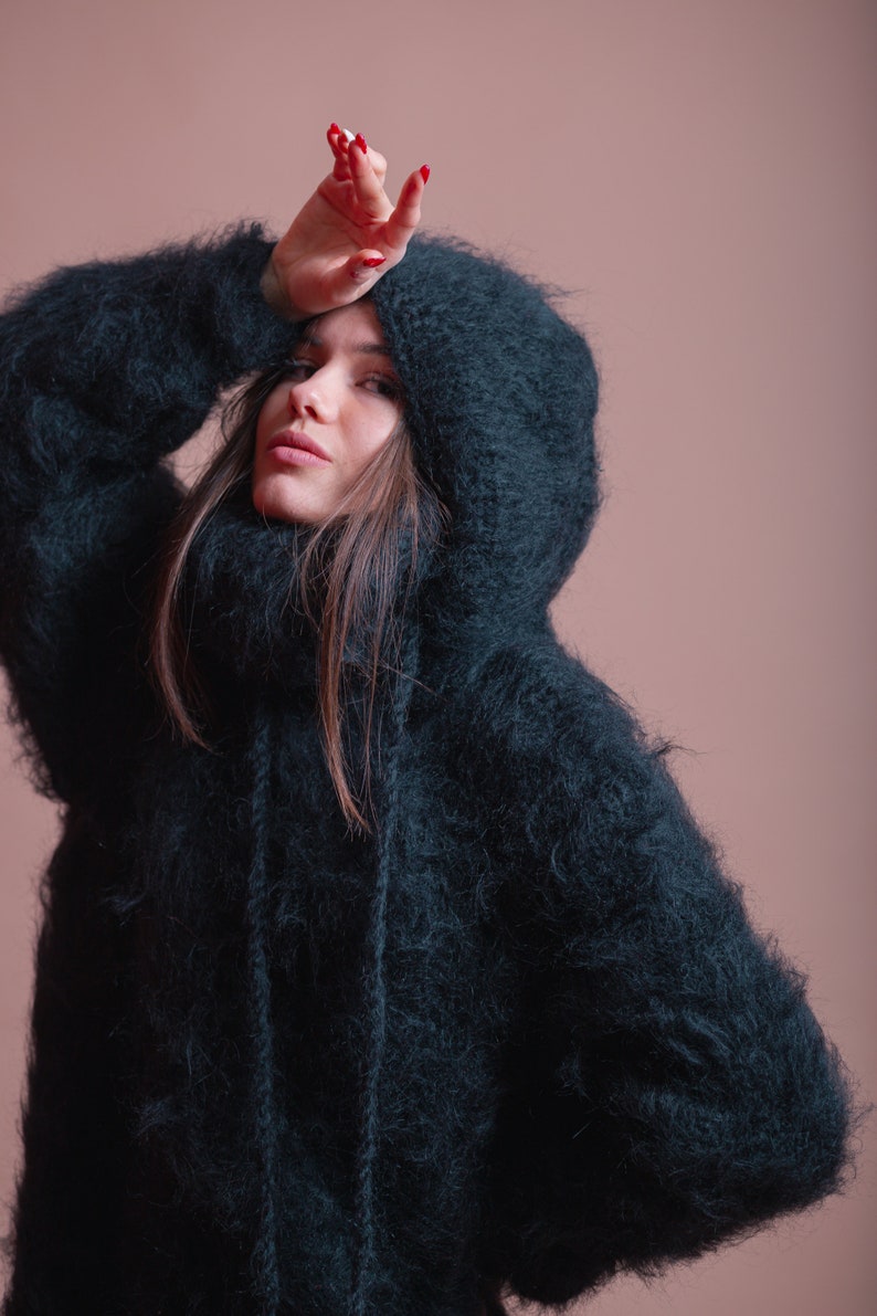 May include: A woman wearing a dark green fuzzy hooded sweater. The sweater has a high neck and a long, loose fit.