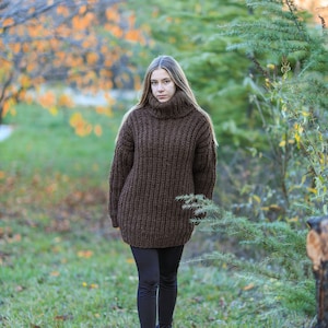 May include: A woman wearing a brown, oversized turtleneck sweater and black leggings walks in a park. The sweater is a chunky knit with a high collar. She is wearing black boots with blue accents. The background features trees and green grass.