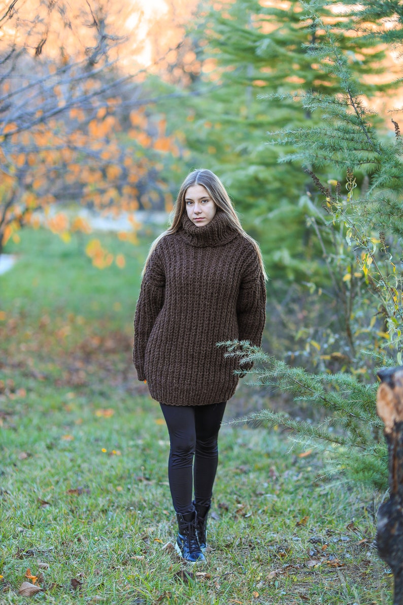 May include: A woman wearing a brown, oversized turtleneck sweater and black leggings walks in a park. The sweater is a chunky knit with a high collar. She is wearing black boots with blue accents. The background features trees and green grass.