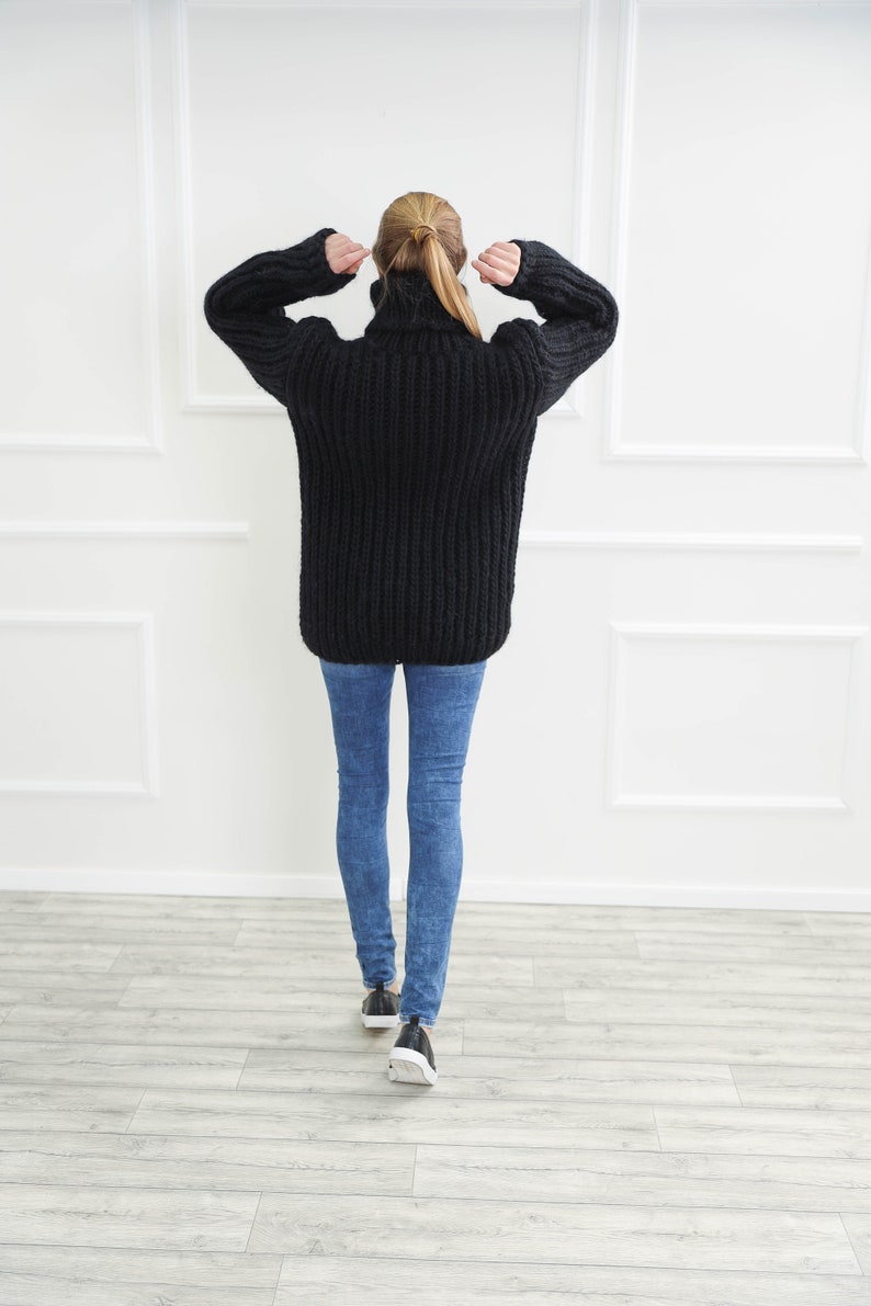 May include: A black, oversized turtleneck sweater with a chunky knit design. The sweater is paired with blue skinny jeans and black sneakers with white soles. The person is facing away from the camera.