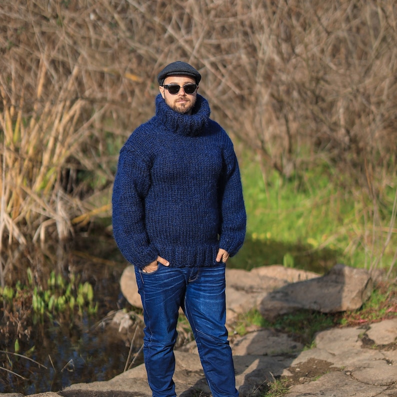 May include: A man wearing a dark blue turtleneck sweater, a dark blue newsboy cap, and dark sunglasses. He is standing in front of a background of brown and green foliage.