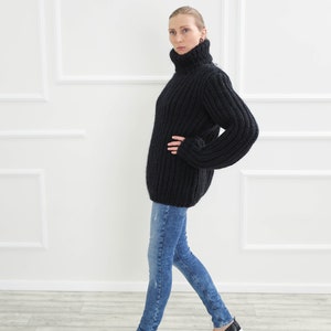 May include: A black, oversized turtleneck sweater with ribbed detailing. The sweater is paired with blue jeans and black slip-on shoes. The model is standing in front of a white wall with decorative molding.