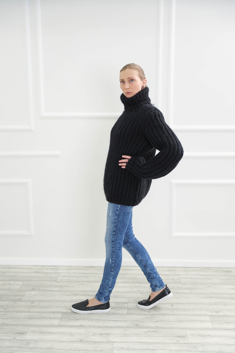 May include: A black, oversized turtleneck sweater with ribbed detailing. The sweater is paired with blue jeans and black slip-on shoes. The model is standing in front of a white wall with decorative molding.