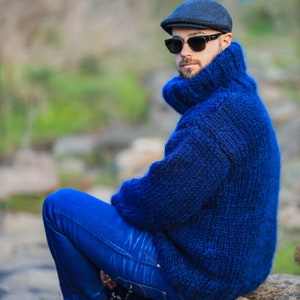 May include: A man wearing a blue knitted turtleneck sweater, blue jeans, and a black cap with sunglasses. He is sitting on a rock outdoors.