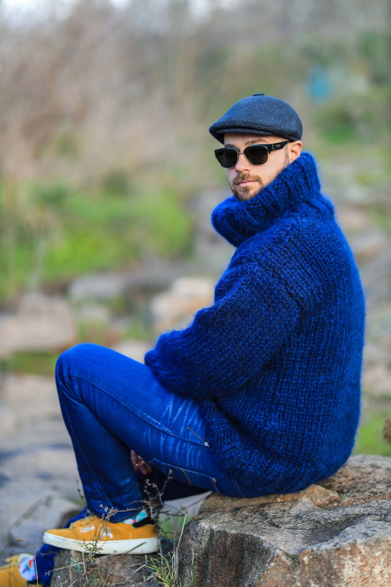 May include: A man wearing a blue knitted turtleneck sweater, blue jeans, and a black cap with sunglasses. He is sitting on a rock outdoors.