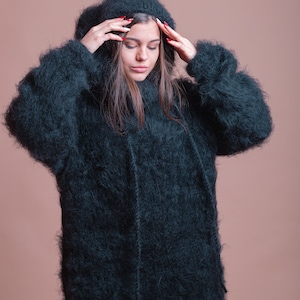 May include: A woman wearing a long, dark green fuzzy sweater with a hood. The sweater is very soft and fluffy.