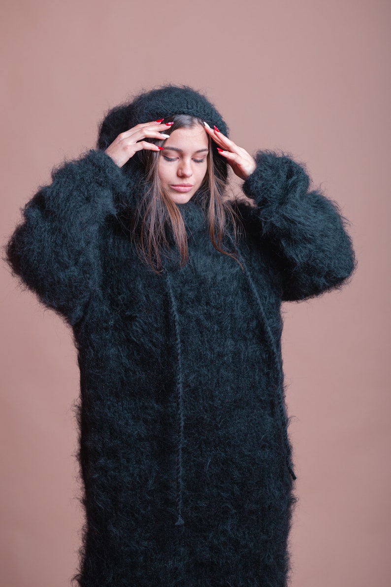 May include: A woman wearing a long, dark green fuzzy sweater with a hood. The sweater is very soft and fluffy.
