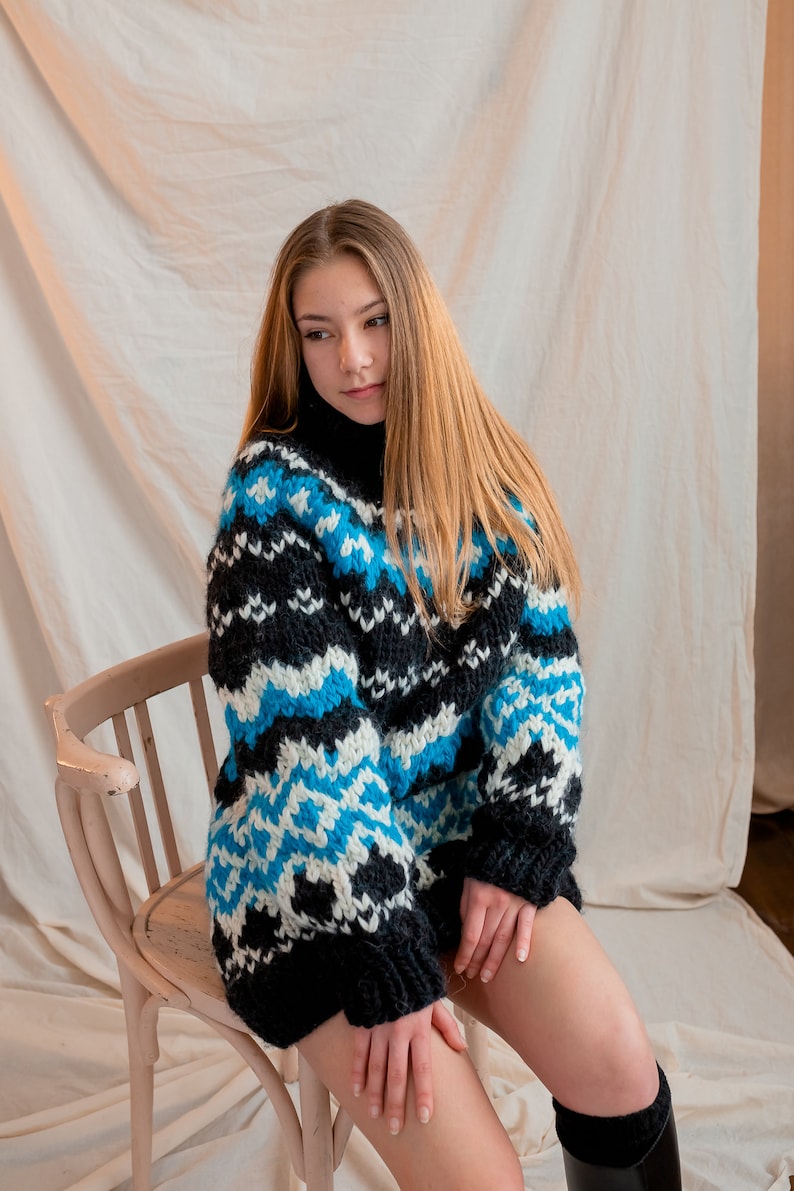 May include: A woman wearing a black, white and blue patterned jumper sits on a wooden chair. The jumper has a high neck and a loose fit.