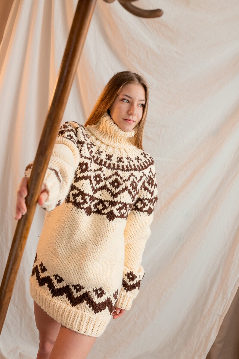 May include: A woman wearing a white and brown knitted sweater with a geometric pattern. The sweater has a turtleneck and long sleeves.