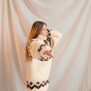 May include: A woman wearing a white and brown knitted sweater with a geometric pattern. The sweater has a turtleneck and long sleeves.