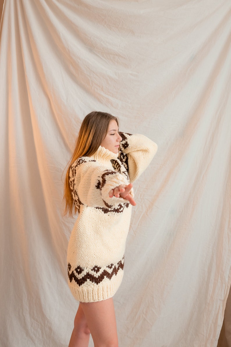 May include: A woman wearing a white and brown knitted sweater with a geometric pattern. The sweater has a turtleneck and long sleeves.