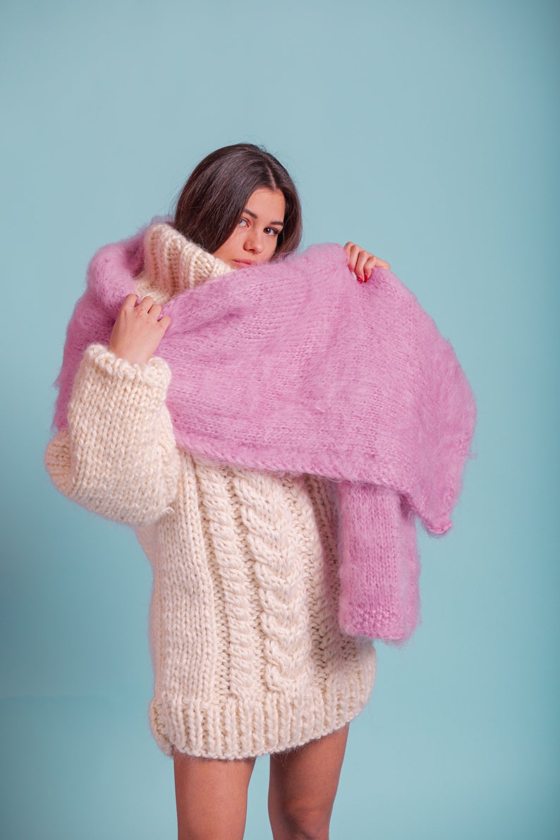 May include: A woman wearing a white cable knit jumper with a pink fluffy scarf draped over her shoulders.