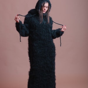 May include: A long, black, fuzzy sweater dress with a hood. The sweater is made of a soft, fluffy material and has a loose fit. The model is wearing the sweater with a pair of black sneakers.