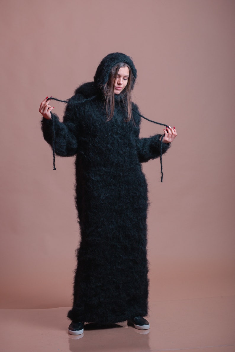 May include: A long, black, fuzzy sweater dress with a hood. The sweater is made of a soft, fluffy material and has a loose fit. The model is wearing the sweater with a pair of black sneakers.