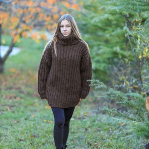 May include: A woman wearing a chunky, dark brown, oversized turtleneck sweater. The sweater has a ribbed knit texture. She is wearing black leggings and black boots. The background shows a blurred outdoor scene with trees and grass.