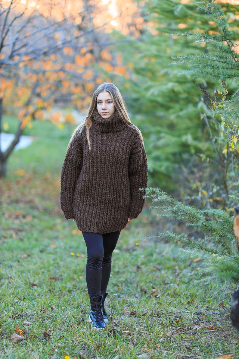 May include: A woman wearing a chunky, dark brown, oversized turtleneck sweater. The sweater has a ribbed knit texture. She is wearing black leggings and black boots. The background shows a blurred outdoor scene with trees and grass.