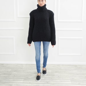 May include: A black, chunky knit turtleneck sweater paired with light blue denim jeans. The sweater has a high, ribbed collar and long sleeves. The jeans are fitted. Black slip-on shoes complete the outfit.