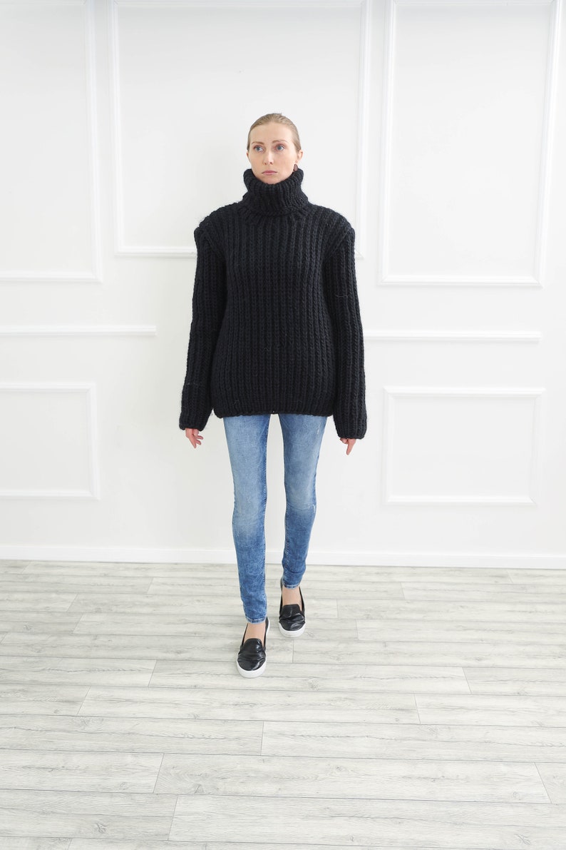 May include: A black, chunky knit turtleneck sweater paired with light blue denim jeans. The sweater has a high, ribbed collar and long sleeves. The jeans are fitted. Black slip-on shoes complete the outfit.