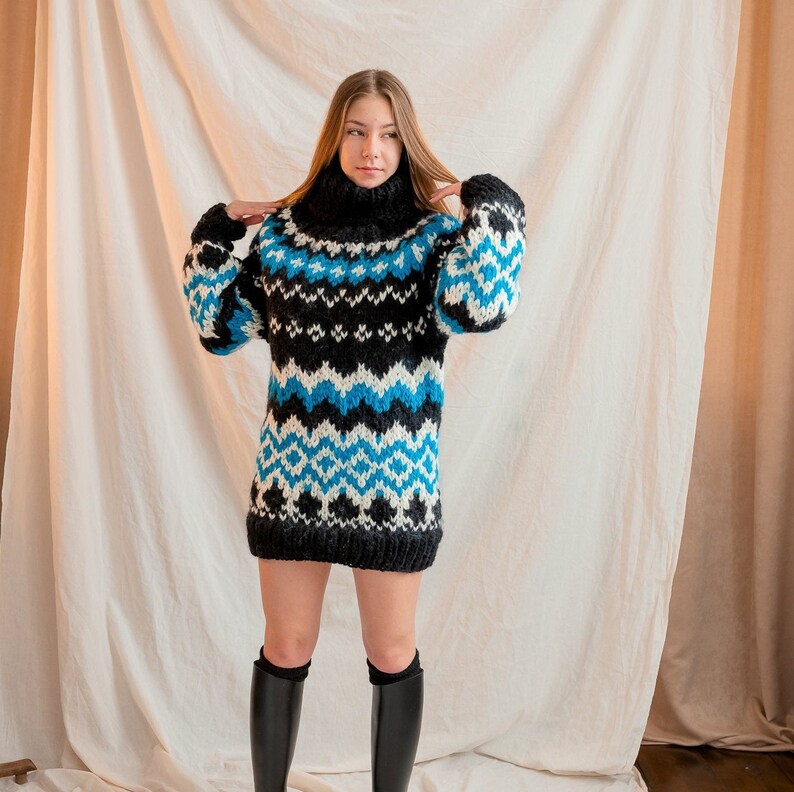 May include: A woman wearing a blue, white and black patterned sweater with a turtleneck. She is standing in front of a white backdrop.