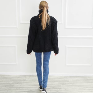 May include: A black, chunky knit turtleneck sweater is worn with blue jeans and black sneakers. The person is facing away from the camera, walking towards a white wall with decorative molding.