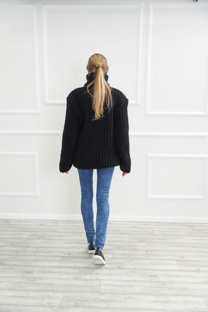 May include: A black, chunky knit turtleneck sweater is worn with blue jeans and black sneakers. The person is facing away from the camera, walking towards a white wall with decorative molding.