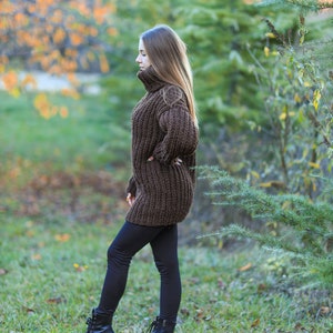May include: A woman wearing a brown, oversized, chunky knit sweater with a turtleneck and black leggings. She is also wearing black sneakers with white soles. The background features green grass, trees, and foliage.