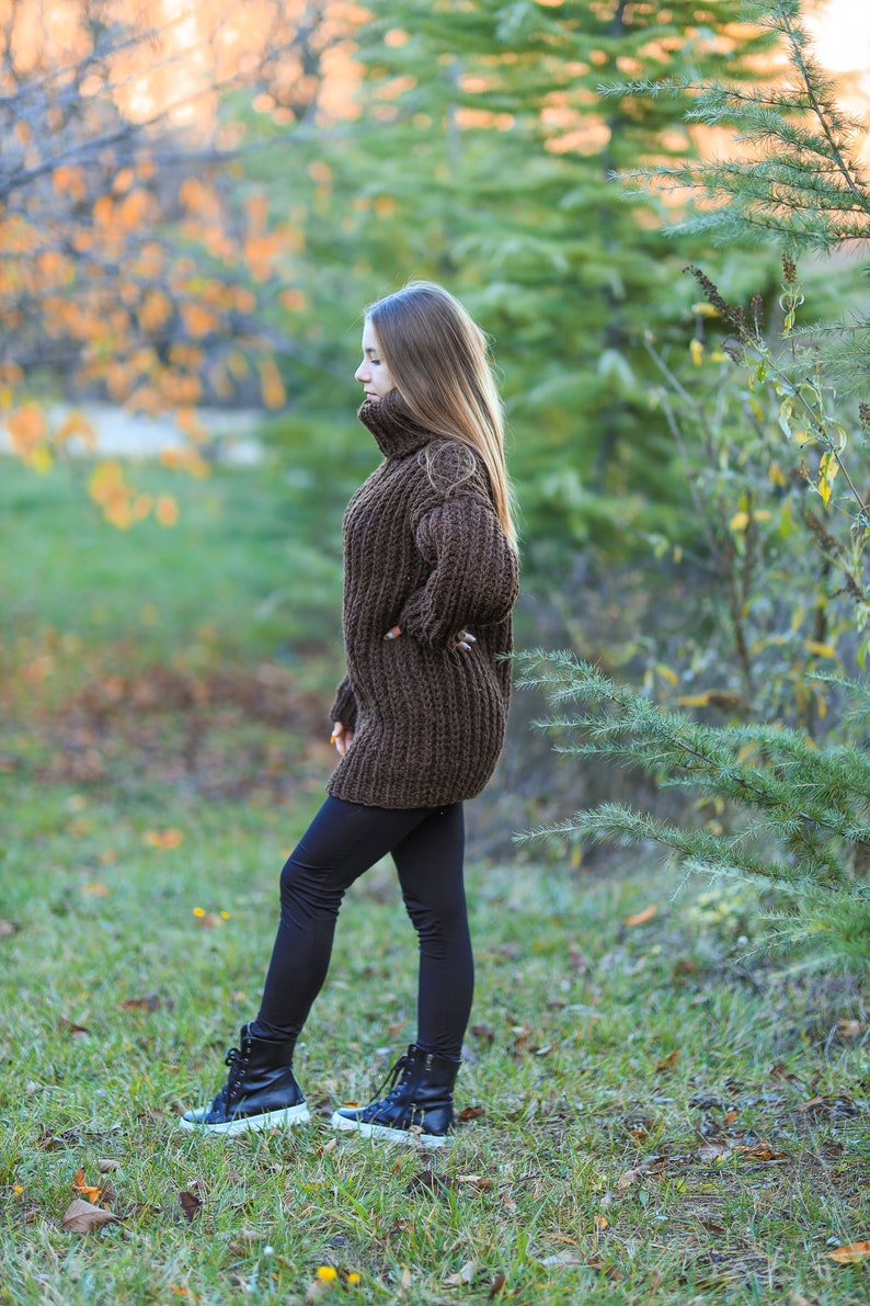 May include: A woman wearing a brown, oversized, chunky knit sweater with a turtleneck and black leggings. She is also wearing black sneakers with white soles. The background features green grass, trees, and foliage.