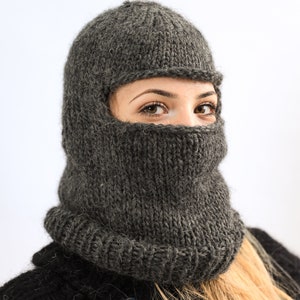Puede incluir: Un pasamontañas de punto gris oscuro con cuello acanalado y cubrecara. El pasamontañas está hecho de un hilo grueso y texturizado y lo lleva una persona con cabello rubio. El pasamontañas cubre la cabeza y el cuello.