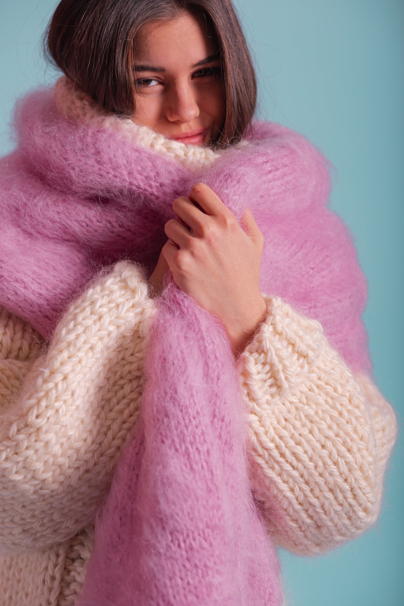 May include: A woman wearing a white and pink knitted jumper with a fluffy pink scarf wrapped around her neck.