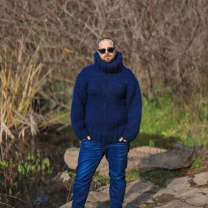 May include: A man wearing a dark blue turtleneck sweater and blue jeans stands in front of a natural background. He is wearing sunglasses and has a beard.