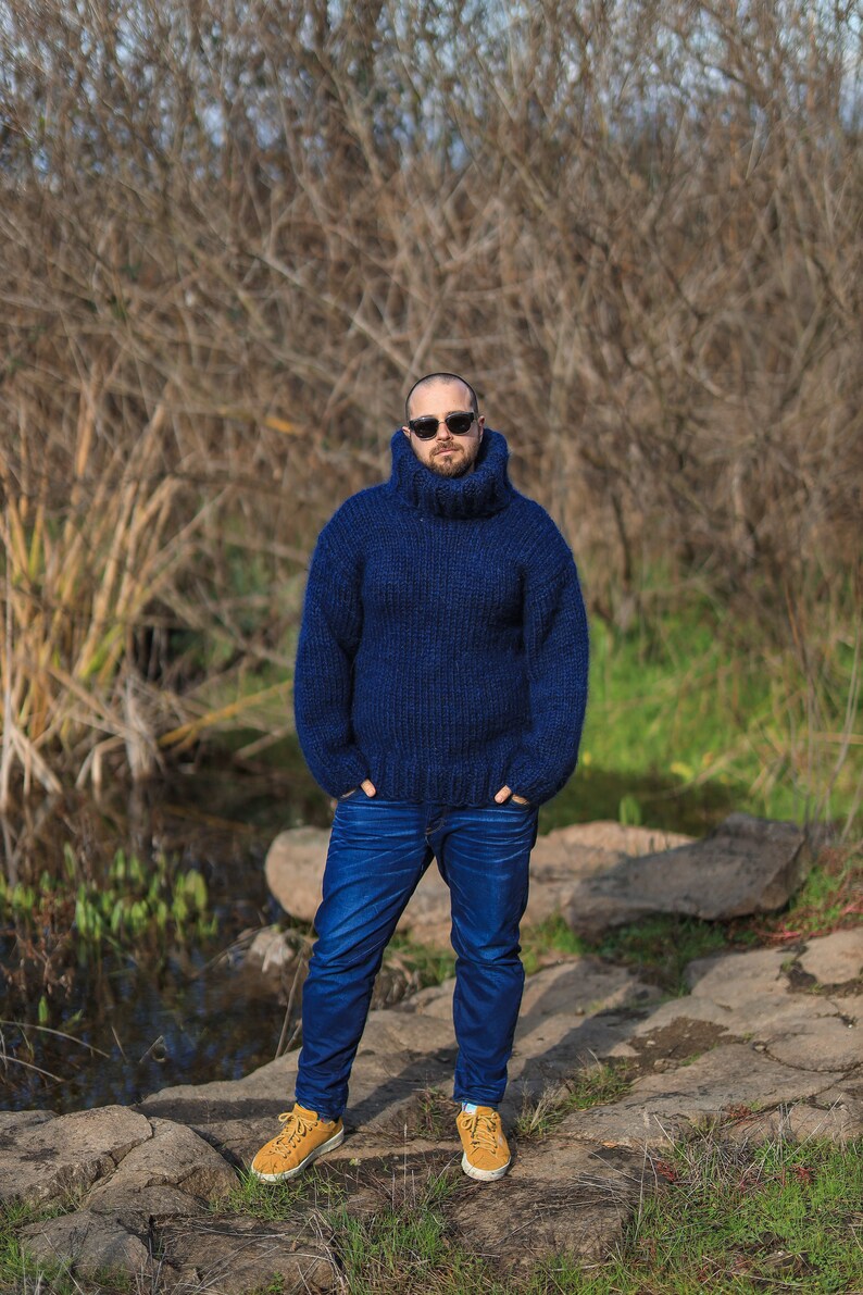 May include: A man wearing a dark blue turtleneck sweater and blue jeans stands in front of a natural background. He is wearing sunglasses and has a beard.