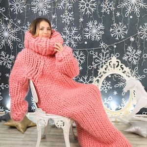 May include: A woman wearing a pink oversized knitted sweater dress with a high turtleneck. The sweater is very thick and chunky. The woman is sitting on a white chair in front of a black background with white snowflakes.