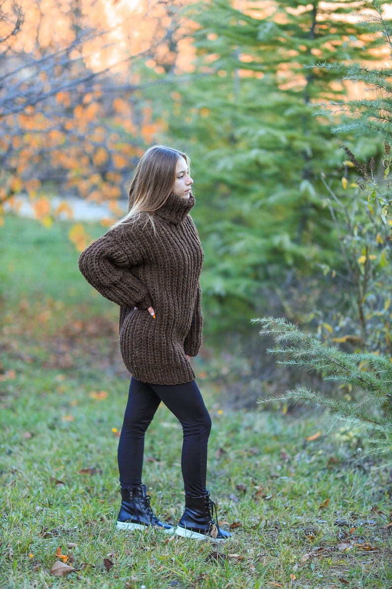 May include: A brown, chunky knit turtleneck sweater dress. The long-sleeved dress is paired with black leggings and black boots with white soles. The model is standing in a grassy area with trees in the background.