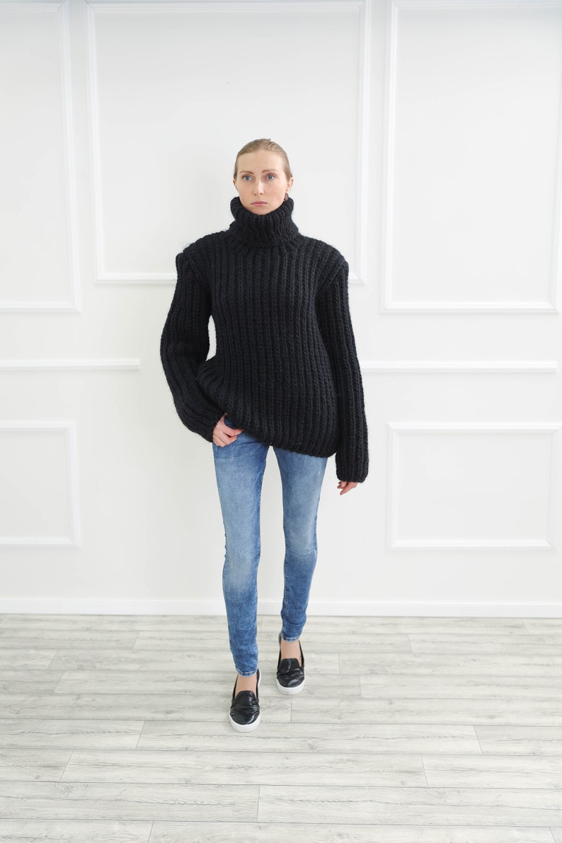 May include: A woman wearing a black, chunky knit turtleneck sweater and blue jeans. The sweater has a high, ribbed collar and a loose fit. The jeans are a light wash, and the woman is wearing black loafers.