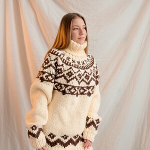 May include: A white and brown knitted sweater with a high neck and a geometric pattern. The sweater is oversized and has long sleeves.