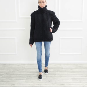 May include: A black, chunky knit turtleneck sweater paired with light blue denim jeans and black loafers. The sweater has a ribbed texture and a high, folded collar. The jeans are fitted, and the loafers have a classic design.