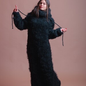 May include: A woman wearing a long, black, fuzzy turtleneck sweater with a high neck. The sweater has long sleeves and is pulled up to her neck. She is standing on a light brown background.