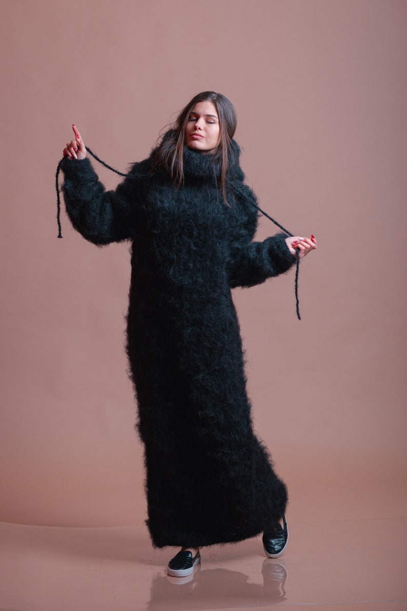 May include: A woman wearing a long, black, fuzzy turtleneck sweater with a high neck. The sweater has long sleeves and is pulled up to her neck. She is standing on a light brown background.
