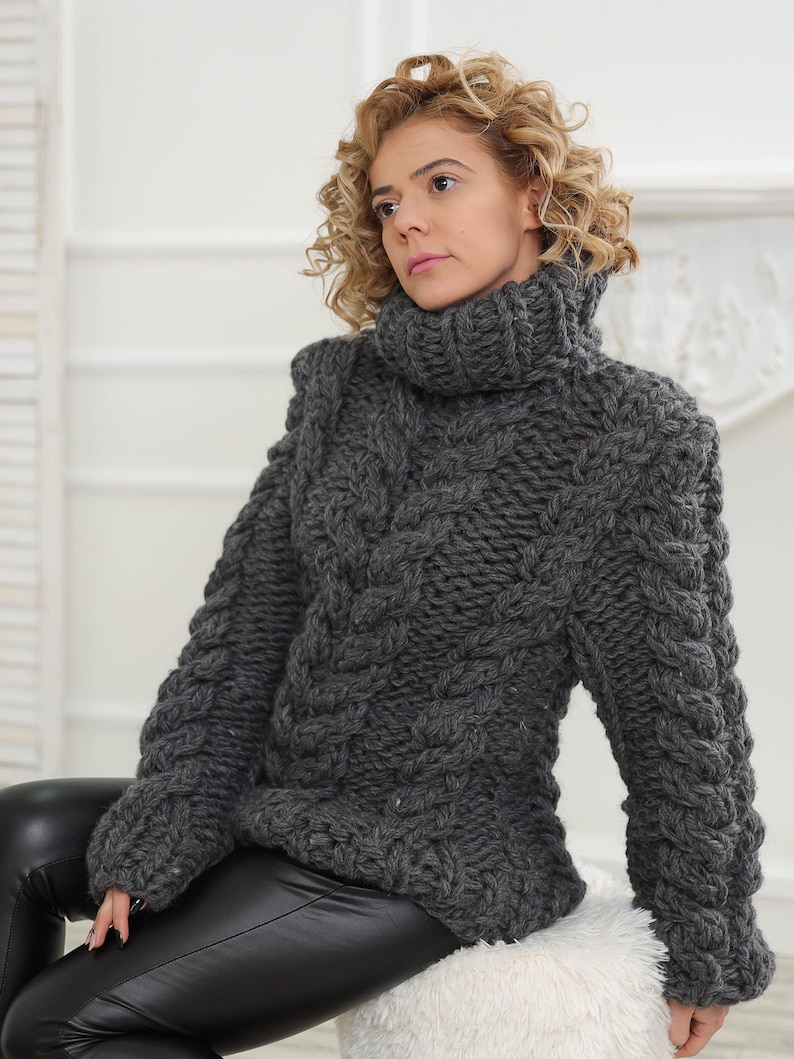 May include: A grey chunky knit turtleneck sweater with a cable knit pattern. The sweater is loose fitting and has long sleeves.